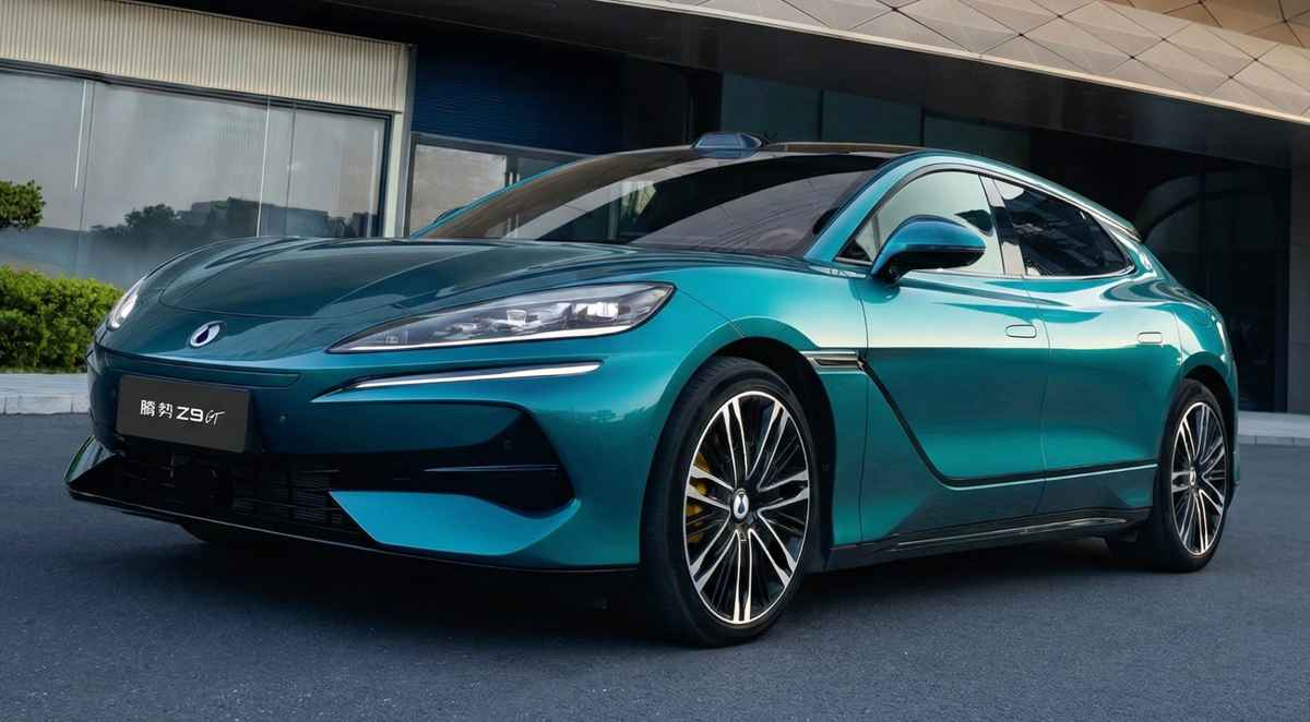 Vista frontal de tres cuartos de un coche deportivo eléctrico Denza Z9 GT de color turquesa metálico brillante, estacionado frente a un edificio de oficinas moderno de vidrio y metal texturizado. La placa de matrícula delantera muestra el texto latino 'Denza Z9 GT' y caracteres chinos (腾势 Z9GT). El vehículo tiene faros delgados y grandes llantas de aleación de varios radios.