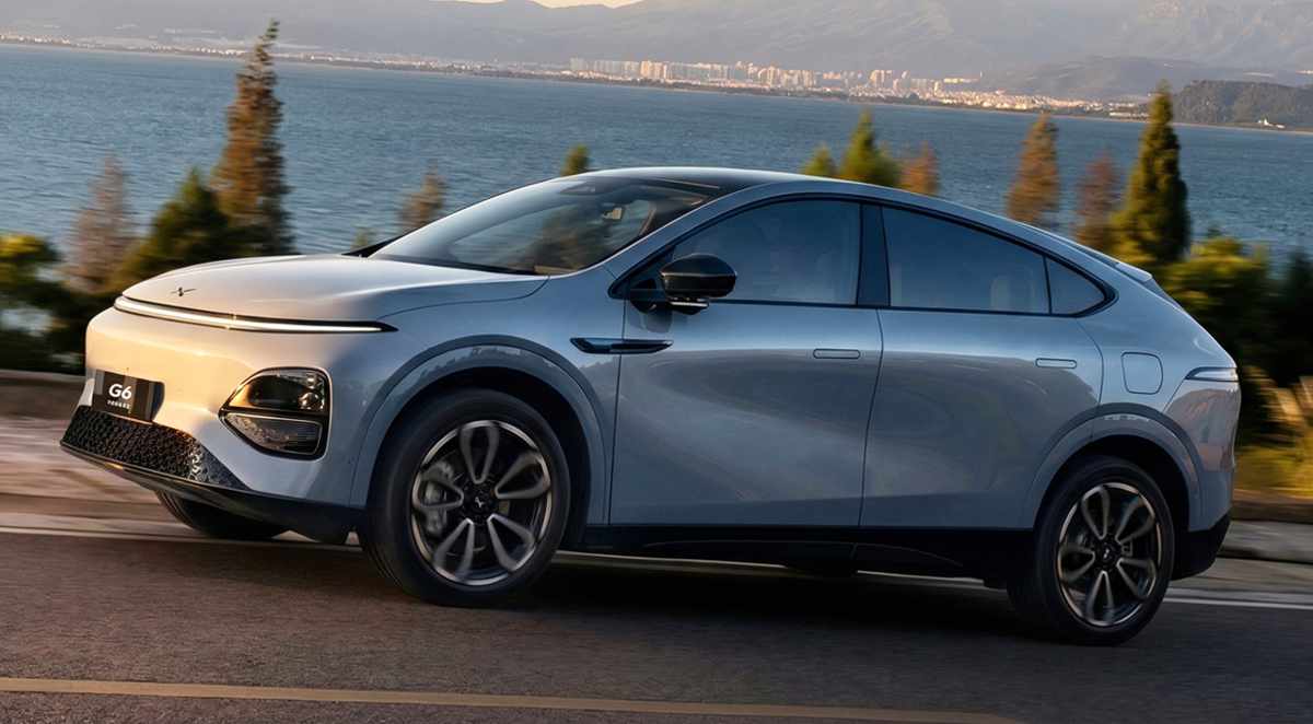 Camioneta SUV coupé Xpeng G6 de color plateado circulando rápidamente sobre una vía asfaltada paralela a un cuerpo de agua. El vehículo oriental exhibe su diseño vanguardista, destacando el frontal sin parrilla, rines bitono y líneas aerodinámicas. El fondo muestra vegetación desenfocada y una ciudad costera bajo un cielo despejado durante el atardecer, transmitiendo movimiento constante.