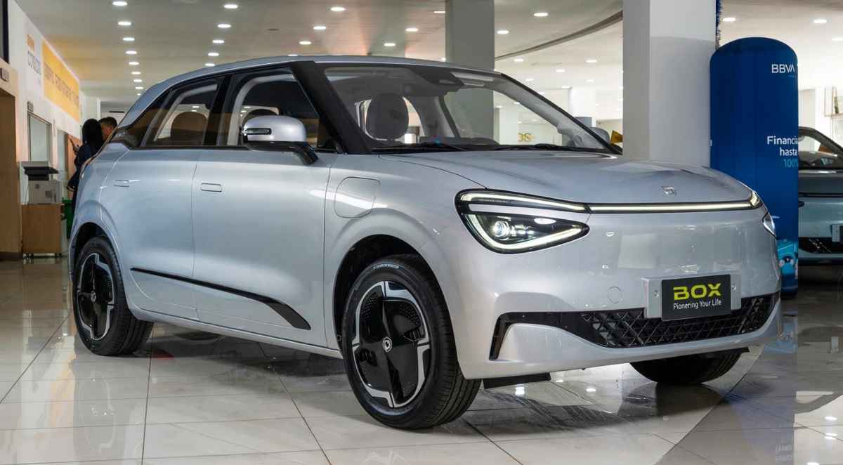 Fotografía en primer plano de tres cuartos delantero de un carro eléctrico Dongfeng Box plateado brillante, estacionado en un showroom de concesionario iluminado en Colombia. Se ven las luces LED encendidas, manijas de puertas enrasadas y rines negros con diseño aerodinámico. El fondo muestra el piso de baldosa reflectante y pancartas publicitarias con texto en español.