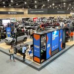Fotografía panorámica desde un ángulo superior del Gran Salón de Corferias durante el Salón del Automóvil de Bogotá. Se observa la inmensa estructura metálica del techo iluminado y la gran escala del recinto ferial, albergando múltiples stands de marcas de carros, vehículos en exhibición y cientos de visitantes recorriendo los amplios pasillos alfombrados de gris.