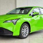 Vista de tres cuartos delantero del sedán GAC Aion ES pintado en color verde reglamentario para taxi eléctrico. El vehículo está exhibido en un concesionario iluminado, mostrando sus faros LED frontales, rines de aluminio y el logo de GAC en el capó.