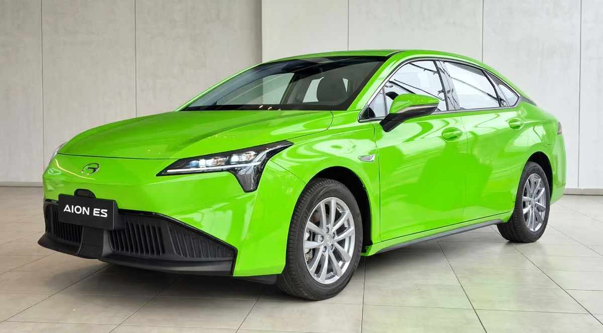 Vista de tres cuartos delantero del sedán GAC Aion ES pintado en color verde reglamentario para taxi eléctrico. El vehículo está exhibido en un concesionario iluminado, mostrando sus faros LED frontales, rines de aluminio y el logo de GAC en el capó.