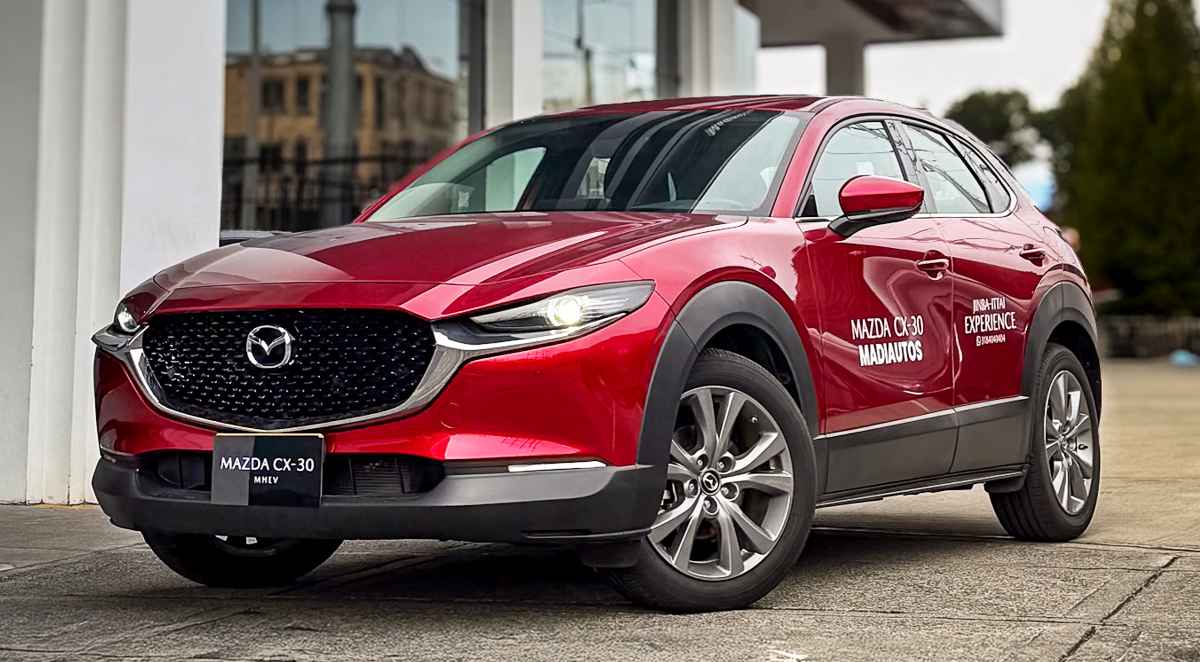 Vista de tres cuartos delantero de una camioneta Mazda CX-30 híbrida de color rojo brillante, parqueada en las afueras del concesionario. En su puerta lateral derecha destacan las calcomanías oficiales de Madiautos, invitando a conocer su portafolio de vehículos sin pico y placa.