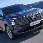 Fotografía en movimiento de una camioneta Changan CS75 Plus de color oscuro circulando por una carretera asfaltada con montañas de fondo. Se destaca su amplia parrilla frontal con diseño paramétrico iluminado, faros LED delgados y rines de aleación, representando la línea de vehículos que adoptará la nueva tecnología híbrida Blue Core de la marca.