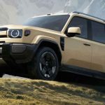 Vista frontal y lateral de la nueva camioneta todoterreno híbrida Chery iCar V27 en color beige, parqueada sobre una colina de pasto con montañas al fondo. Destaca su diseño exterior cuadrado y robusto, faros redondos, rines oscuros de cinco radios y pasos de rueda prominentes en color negro.