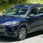 Una vista dinámica de un SUV Toyota Corolla Cross Híbrido de color azul oscuro, la versión actualizada, conduciendo en una carretera de Colombia rodeada de vegetación. Una toma de primer plano muestra la insignia frontal, confirmando su posición como el carro híbrido más vendido en el país en febrero de 2026 (#1).