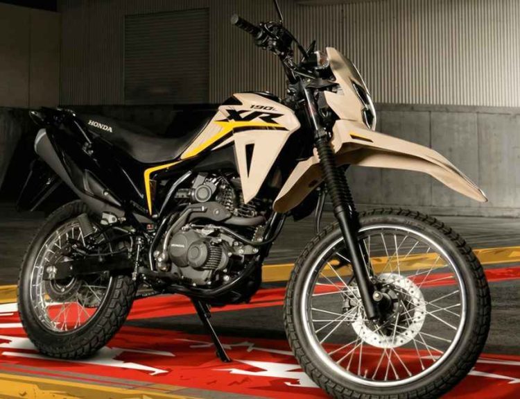 Una vista en ángulo frontal de la motocicleta Honda XR190L de color arena y negro, una versión actualizada, estacionada en un entorno de garaje moderno sobre un suelo con diseño de logo XR, ilustrando su éxito comercial y repunte de ventas del 122,3% en Colombia en febrero de 2026 (#6).