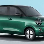 Vista frontal de tres cuartos del nuevo carro eléctrico compacto Wuling MINI EV 2026 color verde perlado con techo negro brillante, mostrando sus renovados faros redondos LED, rines blancos tipo trébol y diseño cuadrado, estacionado en un entorno urbano animado.