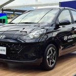 Vista frontal de tres cuartos de un automóvil Hyundai Grand i10 hatchback de color negro, exhibido sobre una tarima de madera dentro de una gran carpa instalada en el estadio El Campín de Bogotá. El vehículo muestra calcomanías de 'Hyundai Patrocinador Oficial FIFA World Cup 2026' en la puerta del conductor. De fondo se aprecian las gradas amarillas y rojas del estadio, césped artificial y otros vehículos Hyundai en exhibición.