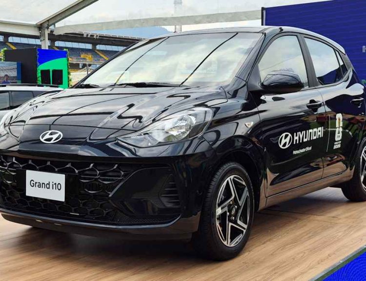 Vista frontal de tres cuartos de un automóvil Hyundai Grand i10 hatchback de color negro, exhibido sobre una tarima de madera dentro de una gran carpa instalada en el estadio El Campín de Bogotá. El vehículo muestra calcomanías de 'Hyundai Patrocinador Oficial FIFA World Cup 2026' en la puerta del conductor. De fondo se aprecian las gradas amarillas y rojas del estadio, césped artificial y otros vehículos Hyundai en exhibición.