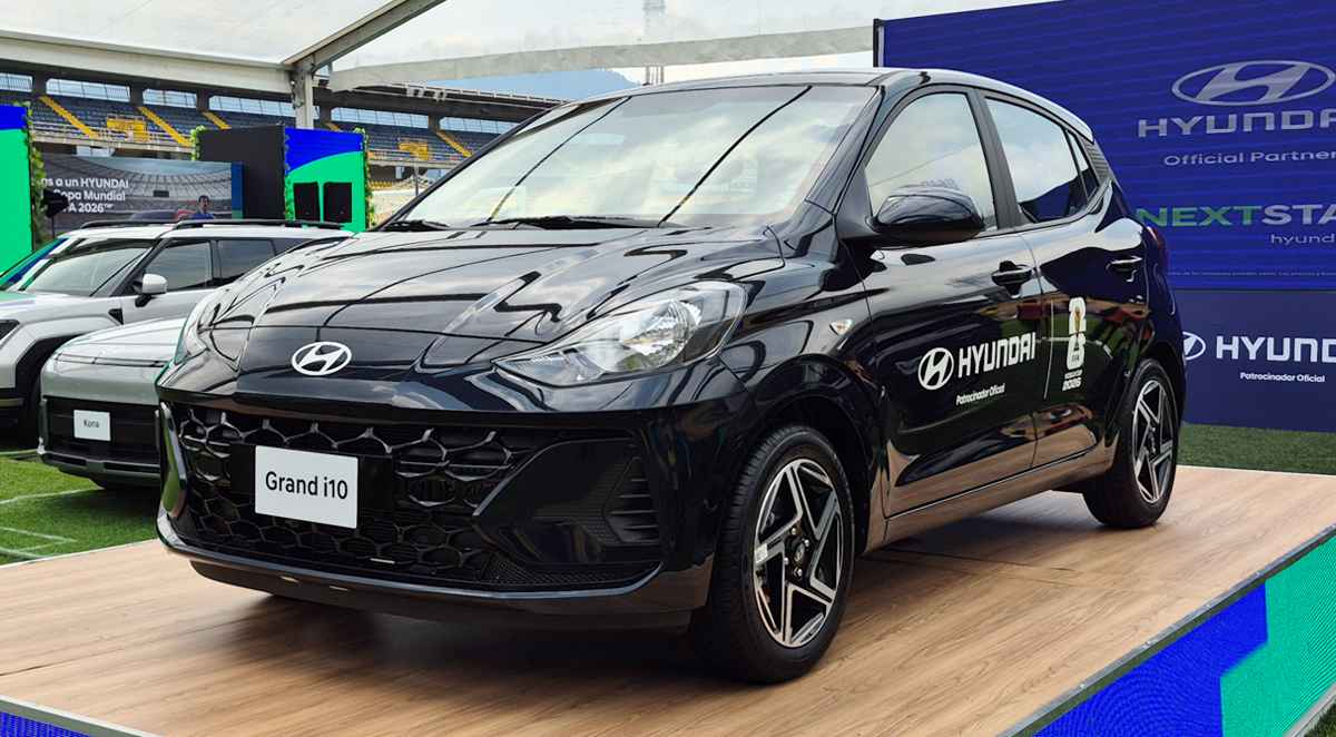 Vista frontal de tres cuartos de un automóvil Hyundai Grand i10 hatchback de color negro, exhibido sobre una tarima de madera dentro de una gran carpa instalada en el estadio El Campín de Bogotá. El vehículo muestra calcomanías de 'Hyundai Patrocinador Oficial FIFA World Cup 2026' en la puerta del conductor. De fondo se aprecian las gradas amarillas y rojas del estadio, césped artificial y otros vehículos Hyundai en exhibición.