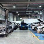 Vista panorámica en profundidad de un moderno y masivo taller de servicio posventa de BYD en Colombia. Se observan múltiples vehículos de la marca, incluyendo SUVs y compactos, estacionados en bahías de mantenimiento numeradas y bajo elevadores hidráulicos. El primer vehículo gris muestra una placa colombiana amarilla (KXR 973), evidenciando la operación local y el amplio respaldo técnico disponible.