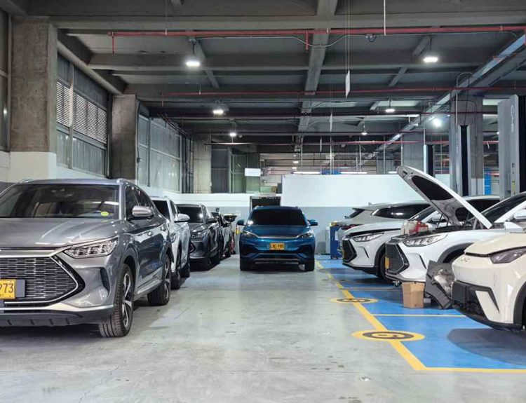 Vista panorámica en profundidad de un moderno y masivo taller de servicio posventa de BYD en Colombia. Se observan múltiples vehículos de la marca, incluyendo SUVs y compactos, estacionados en bahías de mantenimiento numeradas y bajo elevadores hidráulicos. El primer vehículo gris muestra una placa colombiana amarilla (KXR 973), evidenciando la operación local y el amplio respaldo técnico disponible.