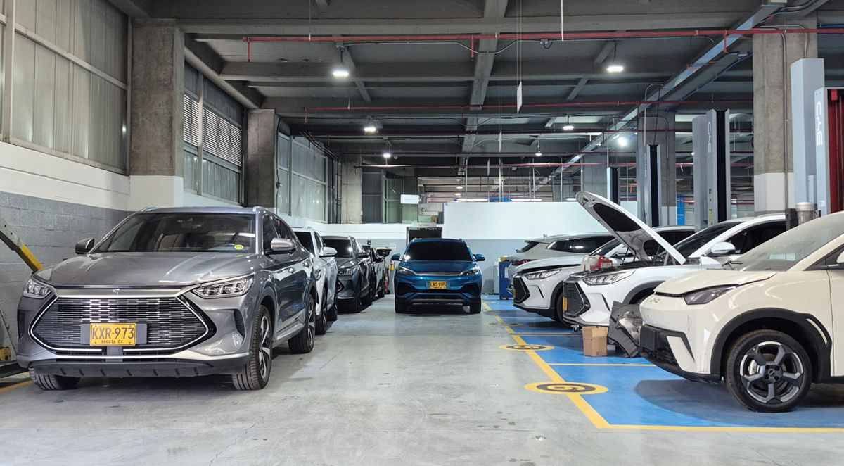Vista panorámica en profundidad de un moderno y masivo taller de servicio posventa de BYD en Colombia. Se observan múltiples vehículos de la marca, incluyendo SUVs y compactos, estacionados en bahías de mantenimiento numeradas y bajo elevadores hidráulicos. El primer vehículo gris muestra una placa colombiana amarilla (KXR 973), evidenciando la operación local y el amplio respaldo técnico disponible.