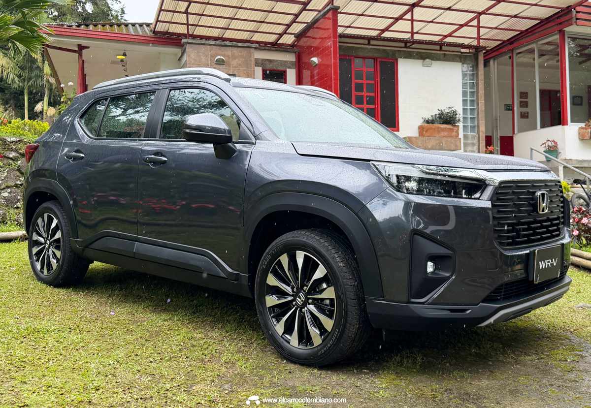 Vista exterior de tres cuartos de la Honda WR-V gris oscura parqueada sobre grama mojada en un paisaje campestre colombiano lluvioso, con el logo de Honda visible en el rine.
