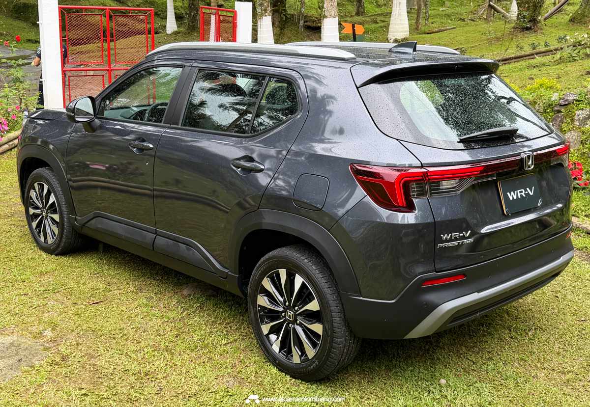 Vista exterior de tres cuartos de la Honda WR-V gris oscura parqueada sobre grama mojada en un paisaje campestre colombiano lluvioso, con el logo de Honda visible en el rine.