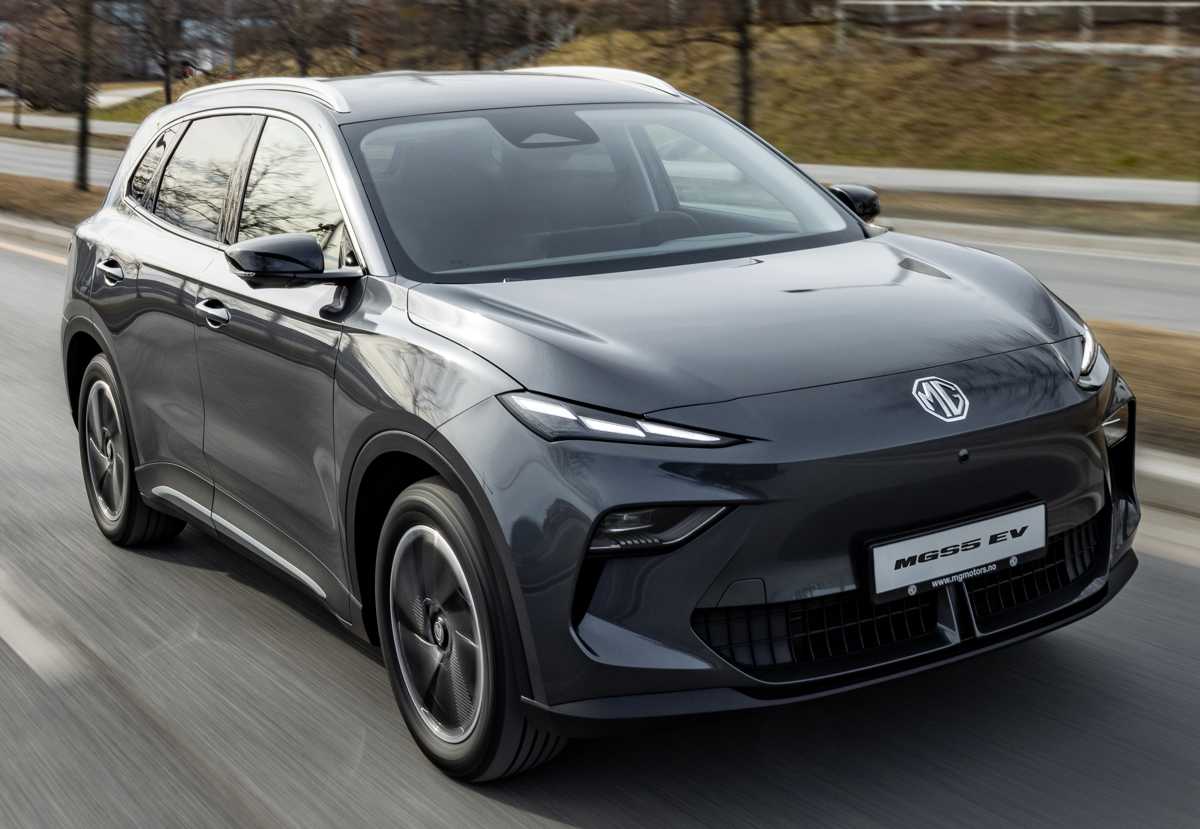Una vista frontal de tres cuartos de un SUV eléctrico MG S5 EV gris oscuro metalizado circulando por una carretera con un fondo borroso de árboles. El elegante vehículo eléctrico tiene una parrilla delantera cerrada y faros LED afilados.