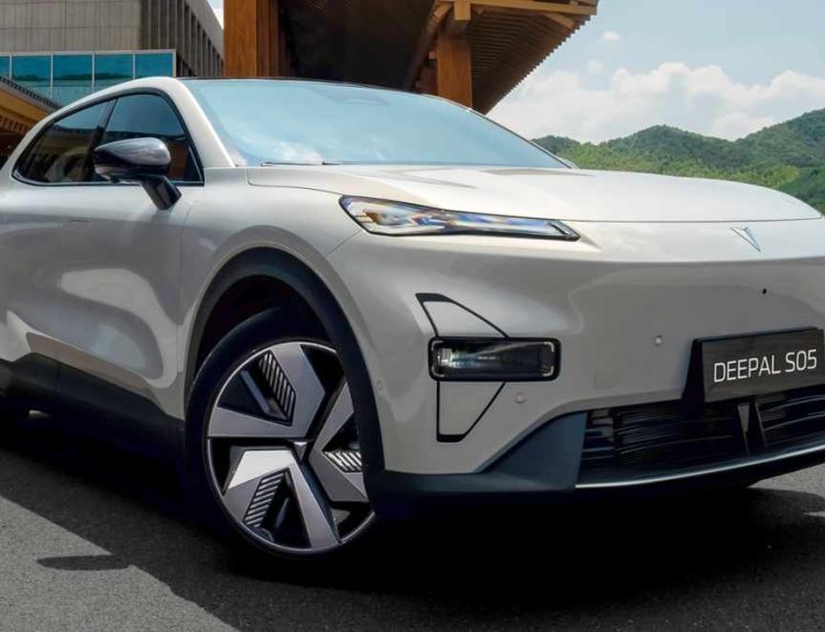 Vista de tres cuartos delantera del SUV Deepal S05 color gris perla, estacionado en una explanada de asfalto frente a un complejo arquitectónico moderno con estructuras de madera. El día está soleado y al fondo se aprecian montañas verdes bajo un cielo azul con nubes dispersas. La placa delantera exhibe el nombre 
