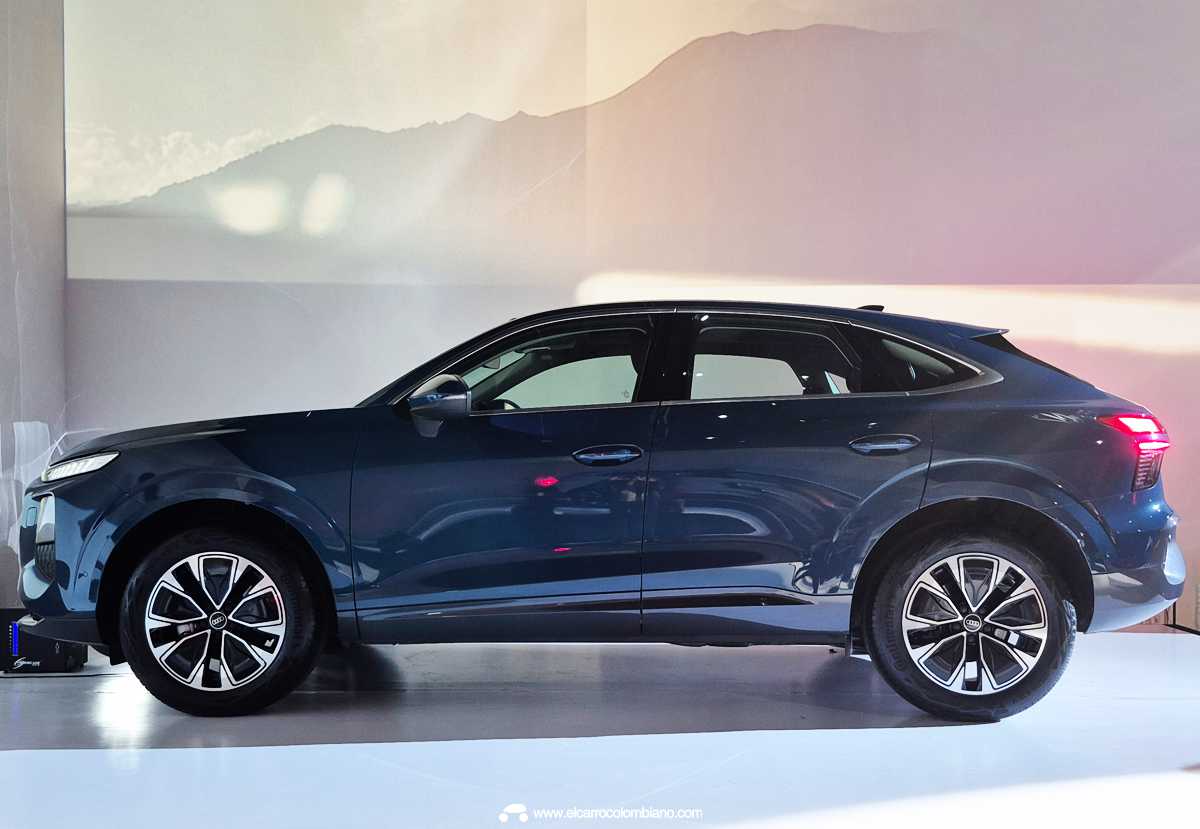 Vista de perfil lateral completo de la nueva Audi Q3 Sportback MHEV color azul oscuro metalizado, exhibida en un estudio de presentación bajo luces de evento sobre un piso blanco. Se aprecia su silueta coupe-SUV con la caída del techo pronunciada, rines de aleación bitono y las luces delanteras y traseras encendidas.