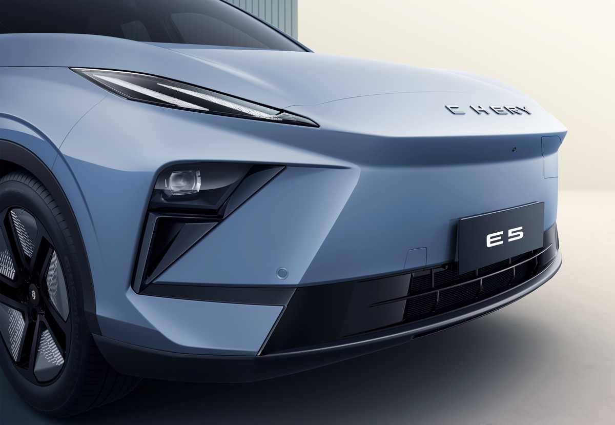 Primer plano detallado de la esquina frontal izquierda de la Chery E5 en color gris plata metalizado. Se enfoca en el diseño afilado del faro LED principal y la moldura negra del parachoques. Parte del rin aerodinámico y el logotipo 'CHERY' son visibles.