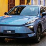 Toma frontal de tres cuartos de la SUV compacta eléctrica Chery E5 en color azul claro metalizado, ubicada en una calle adoquinada del centro histórico colonial de Cartagena, Colombia. El vehículo destaca frente a edificios con fachadas ocre y morado, una gran puerta de madera y balcones, bajo la luz del atardecer. La placa dice 'E5'.