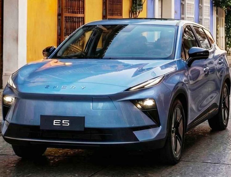 Toma frontal de tres cuartos de la SUV compacta eléctrica Chery E5 en color azul claro metalizado, ubicada en una calle adoquinada del centro histórico colonial de Cartagena, Colombia. El vehículo destaca frente a edificios con fachadas ocre y morado, una gran puerta de madera y balcones, bajo la luz del atardecer. La placa dice 'E5'.