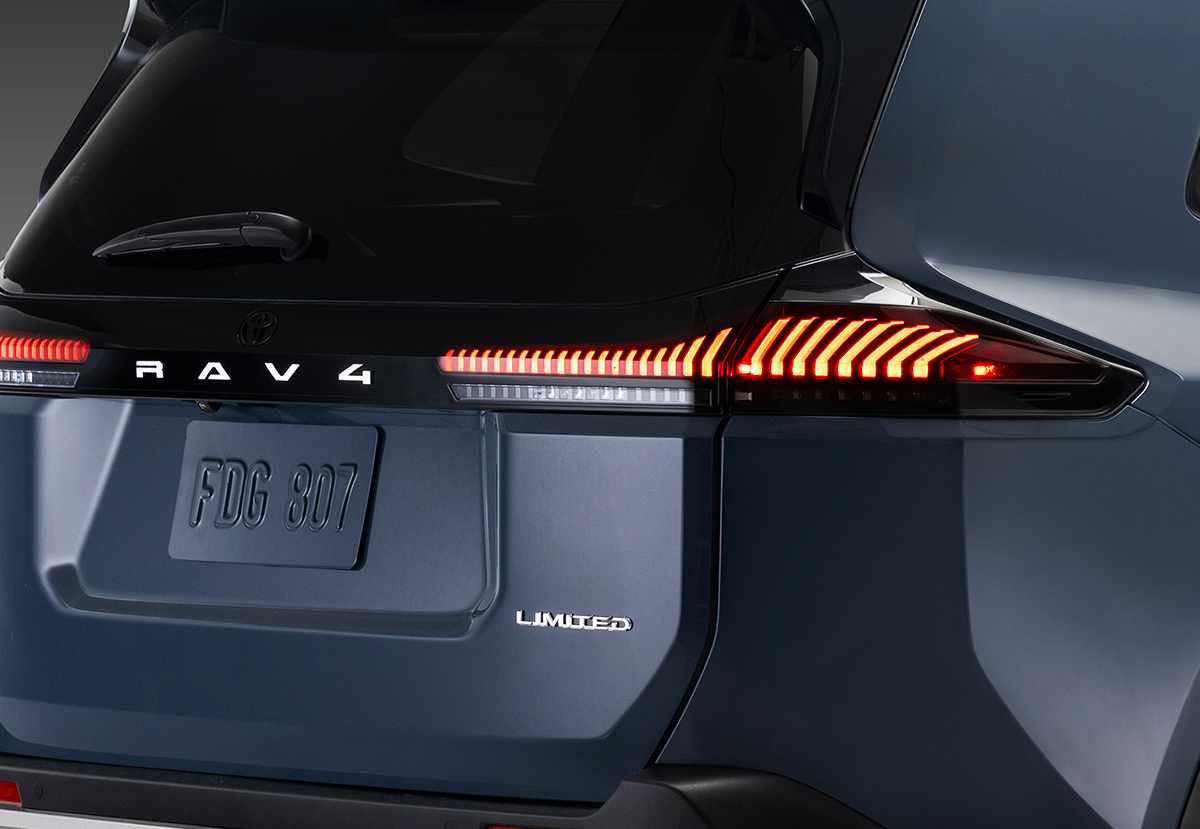 Primer plano detallado de la sección trasera izquierda de una Toyota RAV4 Limited híbrida en color azul oscuro metálico. La imagen se centra en el intrincado diseño de la óptica trasera LED iluminada en rojo brillante, mostrando su compleja firma lumínica. Justo al lado se aprecian con claridad los emblemas cromados 'RAV4' en la moldura central y el distintivo de versión 'LIMITED' debajo. Se destacan la pintura brillante y la zona de la matrícula con el portón trasero.