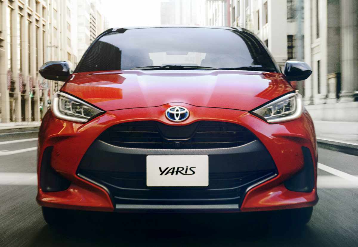 Primer plano frontal de un Toyota Yaris híbrido rojo (versión global) circulando por una calle de ciudad, destacando su parrilla con el emblema 'YARIS' y faros LED.