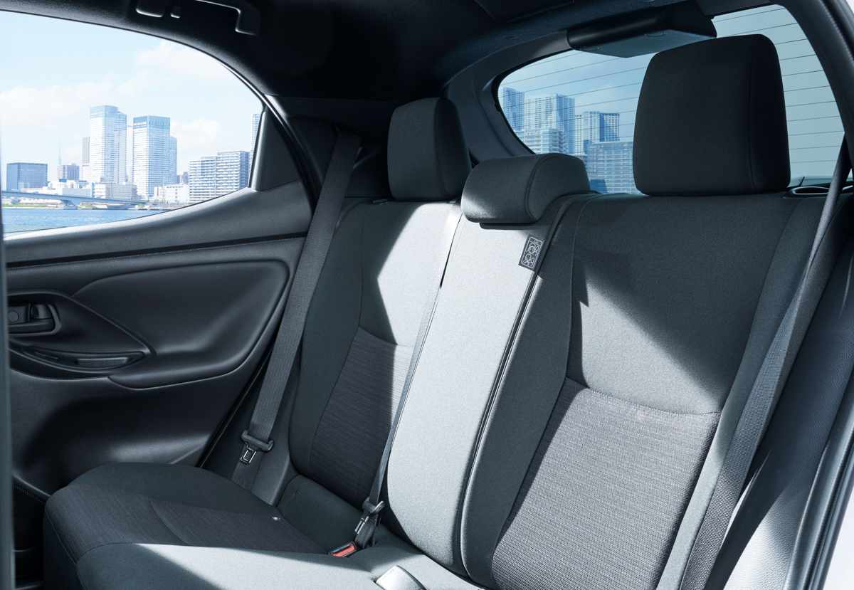 Vista del interior trasero de un Toyota Yaris, mostrando los asientos de tela con patrón, los reposacabezas y una vista panorámica borrosa de un horizonte urbano frente al agua a través de las ventanas.