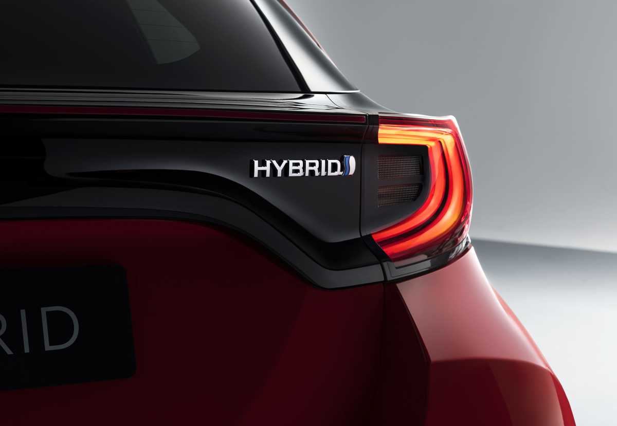 Primer plano detallado de la parte trasera de una Toyota Yaris Híbrida 2026 en color rojo metalizado, mostrando el emblema cromado 'HYBRID' con el característico aro azul de la tecnología Full Hybrid de la marca sobre la moldura negra brillante. Al lado, se aprecia el diseño de la óptica trasera LED encendida.