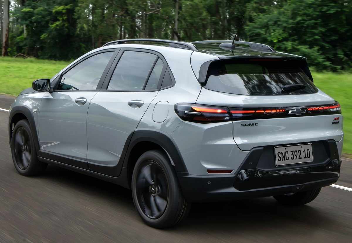 Vista tres cuartos trasera del Chevrolet Sonic 2027 plateado circulando sobre asfalto, resaltando su silueta tipo cupé, la firma lumínica LED horizontal que cruza el baúl, el corbatín negro y los emblemas 