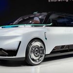 Vista de tres cuartos delantera del concept car Geely Eva Cab en su stand de exhibición en Pekín, mostrando su diseño de minivan blanca con líneas limpias, rines cromados, el parachoques inferior con la inscripción 'ROBOTAXI' y detalles de iluminación LED verde azulado.