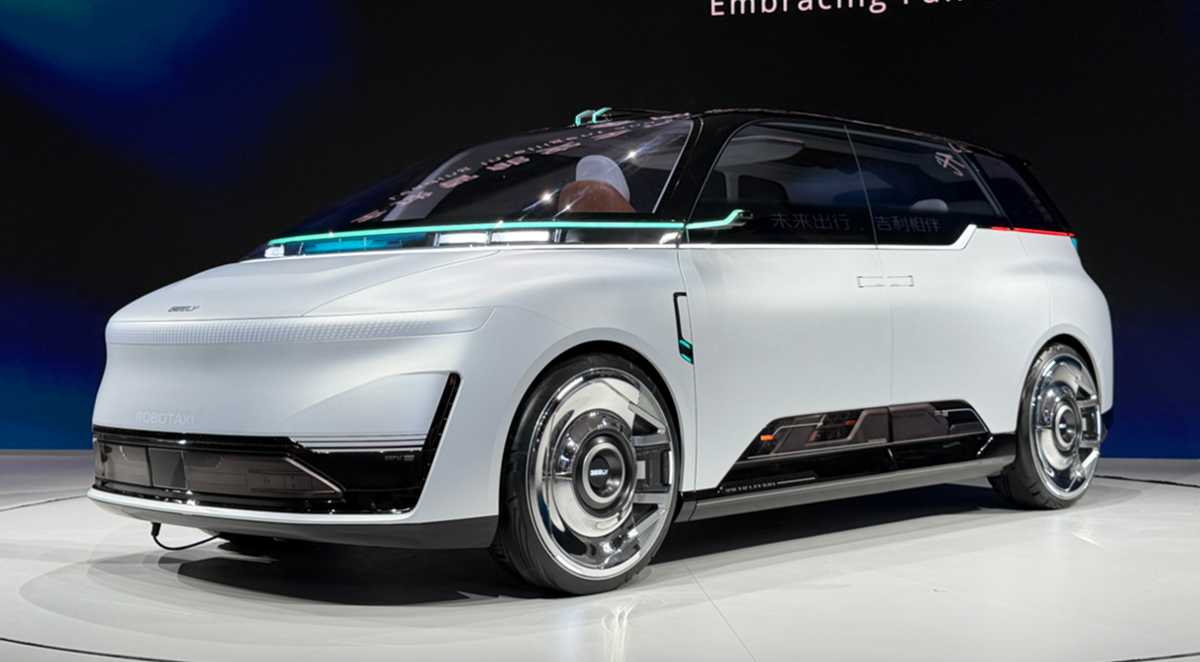 Vista de tres cuartos delantera del concept car Geely Eva Cab en su stand de exhibición en Pekín, mostrando su diseño de minivan blanca con líneas limpias, rines cromados, el parachoques inferior con la inscripción 'ROBOTAXI' y detalles de iluminación LED verde azulado.