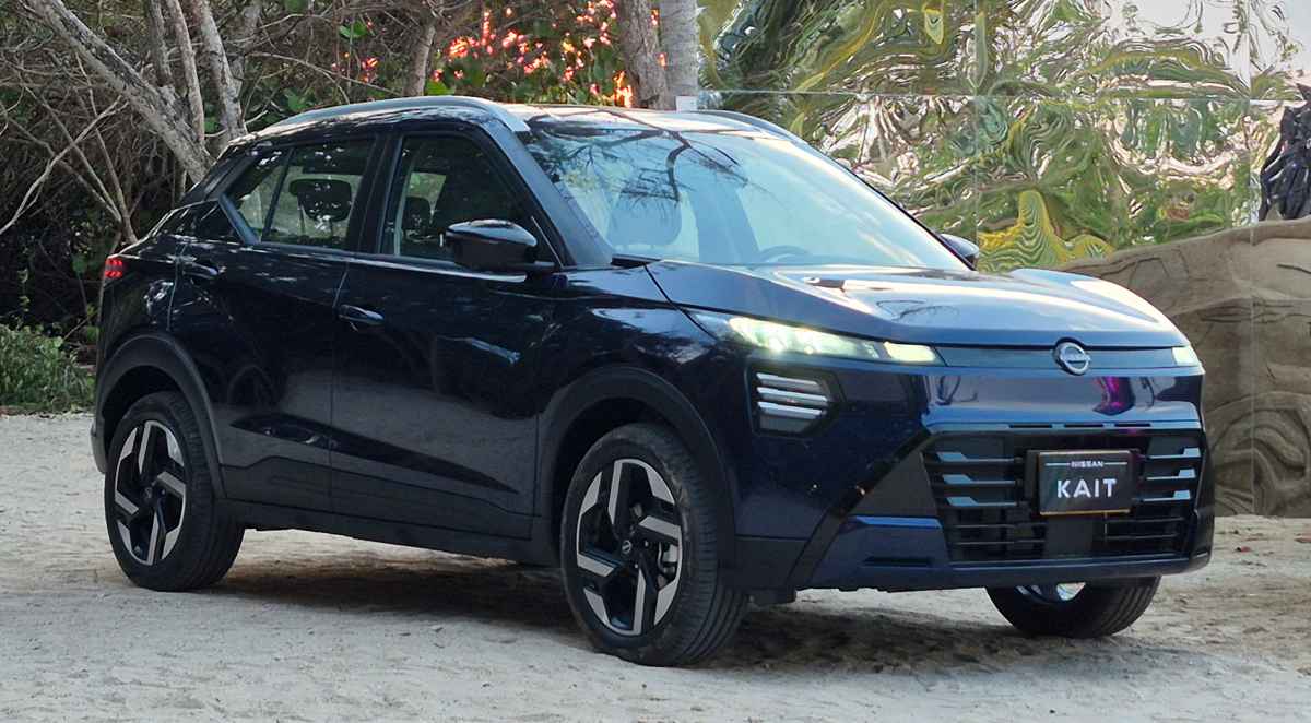 Vista frontal de tres cuartos de la nueva SUV Nissan Kait en color azul oscuro, estacionada sobre una superficie de arena. Destaca su moderna parrilla frontal con detalles horizontales, faros LED encendidos y rines bitono, frente a un fondo con árboles y una estructura reflectante.