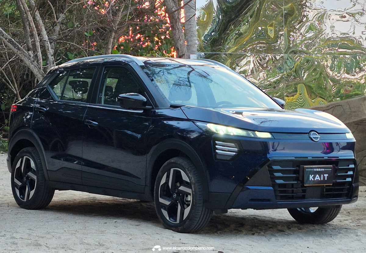 Vista frontal de tres cuartos de la nueva SUV Nissan Kait en color azul oscuro, estacionada sobre una superficie de arena. Destaca su moderna parrilla frontal con detalles horizontales, faros LED encendidos y rines bitono, frente a un fondo con árboles y una estructura reflectante.