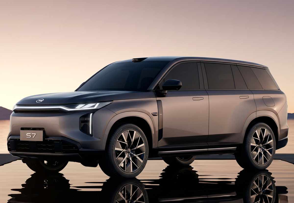 Vista frontal de tres cuartos de la gran SUV híbrida enchufable GAC S7 en color gris mate. Destaca su diseño robusto, rines bitono, carrocería de líneas rectas y una firma lumínica frontal de luces LED delgadas, posando sobre una superficie reflectante al atardecer.