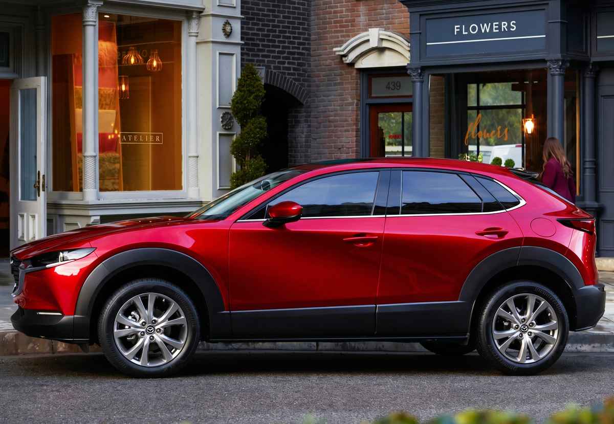 Un SUV Mazda CX-30 rojo brillante estacionado en una calle urbana de aspecto elegante, frente a storefronts con letreros que dicen 