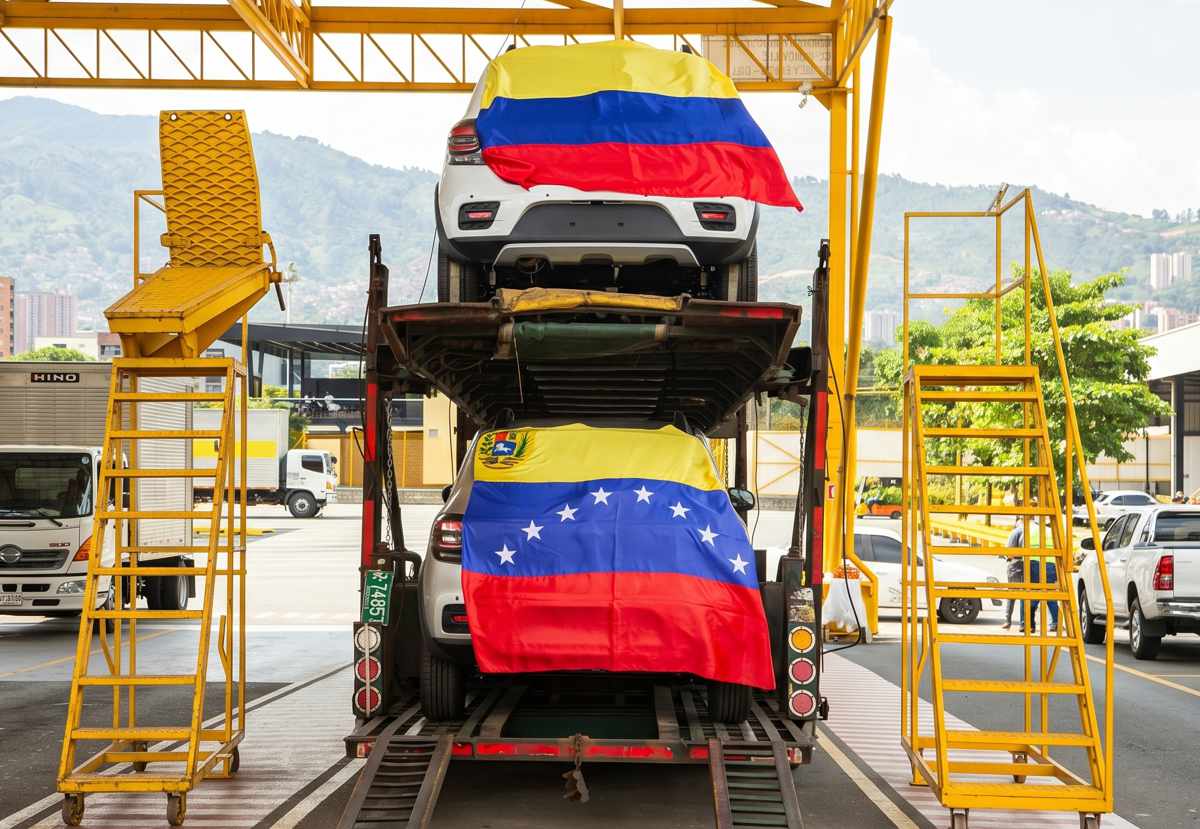 Vehículos blancos recién ensamblados por Renault-Sofasa, cubiertos con las banderas de Colombia y Venezuela, acomodados sobre un camión transportador amarillo tipo niñera, listos para su proceso de exportación.