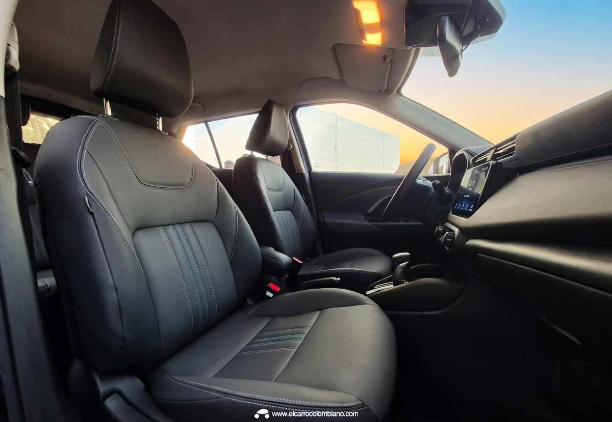 Vista interior lateral de los asientos delanteros de la Nissan Kait, mostrando la cojinería en tela negra con costuras grises, el panel de la puerta y el tablero, con la vista del atardecer a través de la ventana del pasajero.