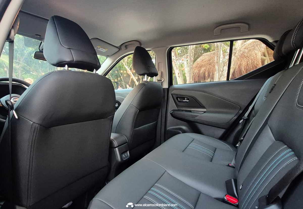 Vista interior desde la puerta trasera abierta de la Nissan Kait, mostrando la banca de asientos posterior en tela negra con costuras grises y la parte trasera de los asientos delanteros, en un entorno de evento de playa.