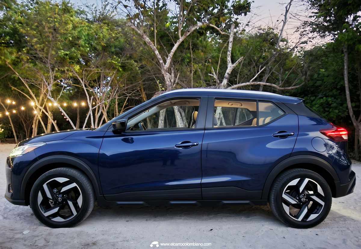 Vista de perfil completo (lateral) de la SUV Nissan Kait en color azul oscuro, estacionada sobre la arena de una playa durante el atardecer, con árboles decorados con luces al fondo.