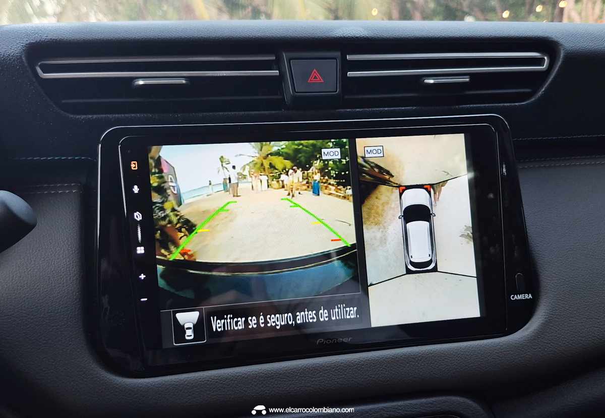 Primer plano de la pantalla táctil central de la Nissan Kait mostrando el funcionamiento del sistema de cámara de visión periférica 360 grados, con visualización dividida de la vista trasera y cenital del vehículo en un entorno de playa.