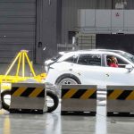 Vista lateral del momento exacto de una prueba de choque en el Centro de Seguridad de Geely. Una plataforma móvil amarilla impacta violentamente la parte trasera de una camioneta Zeekr 7X color blanco, con un maniquí de pruebas visible al volante.