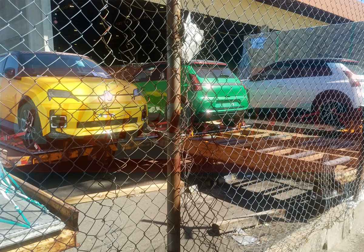 Fotografía espía tomada a través de una malla metálica que muestra tres unidades del Renault 5 eléctrico estacionadas sobre remolques de carga. Destaca frontalmente una unidad de color amarillo encendido, revelando sus característicos faros cuadrados y diseño retro.