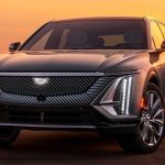 Fotografía frontal de la camioneta eléctrica Cadillac Lyriq en un paisaje desértico al atardecer. Destaca su imponente parrilla iluminada con patrones diagonales, el tradicional escudo de la marca y sus estilizados faros LED verticales.