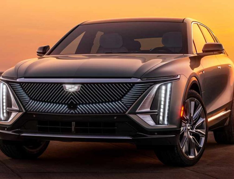 Fotografía frontal de la camioneta eléctrica Cadillac Lyriq en un paisaje desértico al atardecer. Destaca su imponente parrilla iluminada con patrones diagonales, el tradicional escudo de la marca y sus estilizados faros LED verticales.