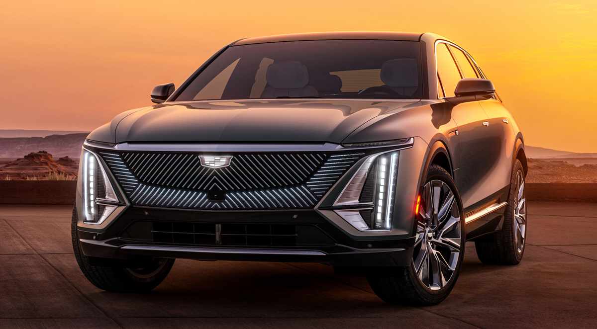 Fotografía frontal de la camioneta eléctrica Cadillac Lyriq en un paisaje desértico al atardecer. Destaca su imponente parrilla iluminada con patrones diagonales, el tradicional escudo de la marca y sus estilizados faros LED verticales.