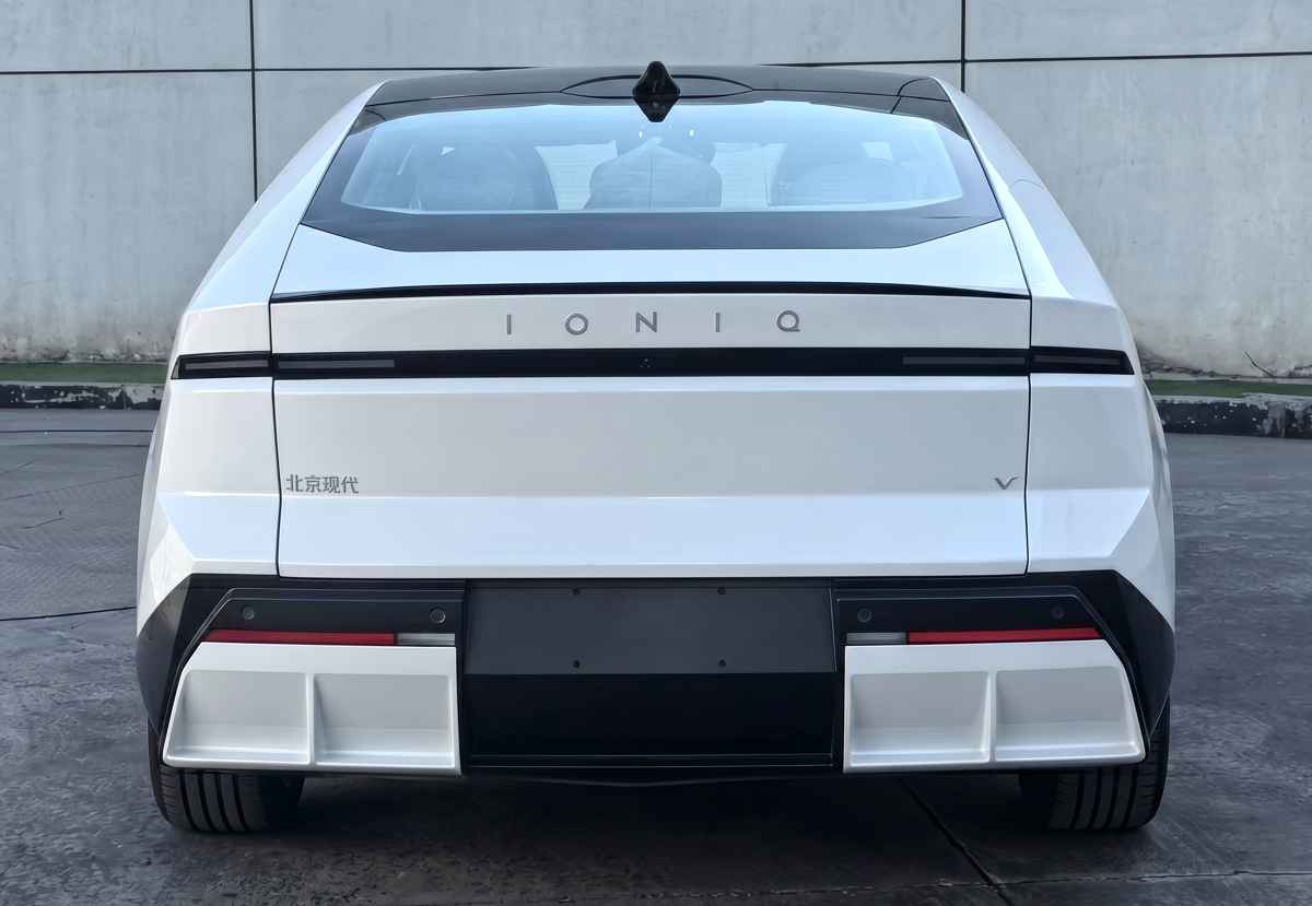 Vista posterior de homologación del sedán Hyundai Ioniq V en color blanco. Resalta una franja de luces traseras oscura que cruza a lo ancho, la inscripción IONIQ en el centro de la compuerta y un difusor inferior aerodinámico prominente.