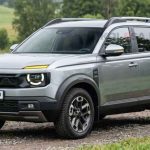Proyección fotorrealista del nuevo Lada Niva 2028 en color gris plata, rodando sobre un camino de tierra, destacando su frontal retro con faros redondos LED y letras integradas en la parrilla.