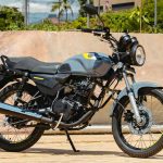Motocicleta AKT AK125 NKD color gris nardo con detalles amarillos estacionada de perfil sobre una acera urbana, mostrando su diseño clásico con faro redondo y rines de radios.